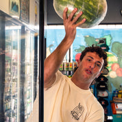Person holding a large watermelon in front of a refrigerator with drinks inside