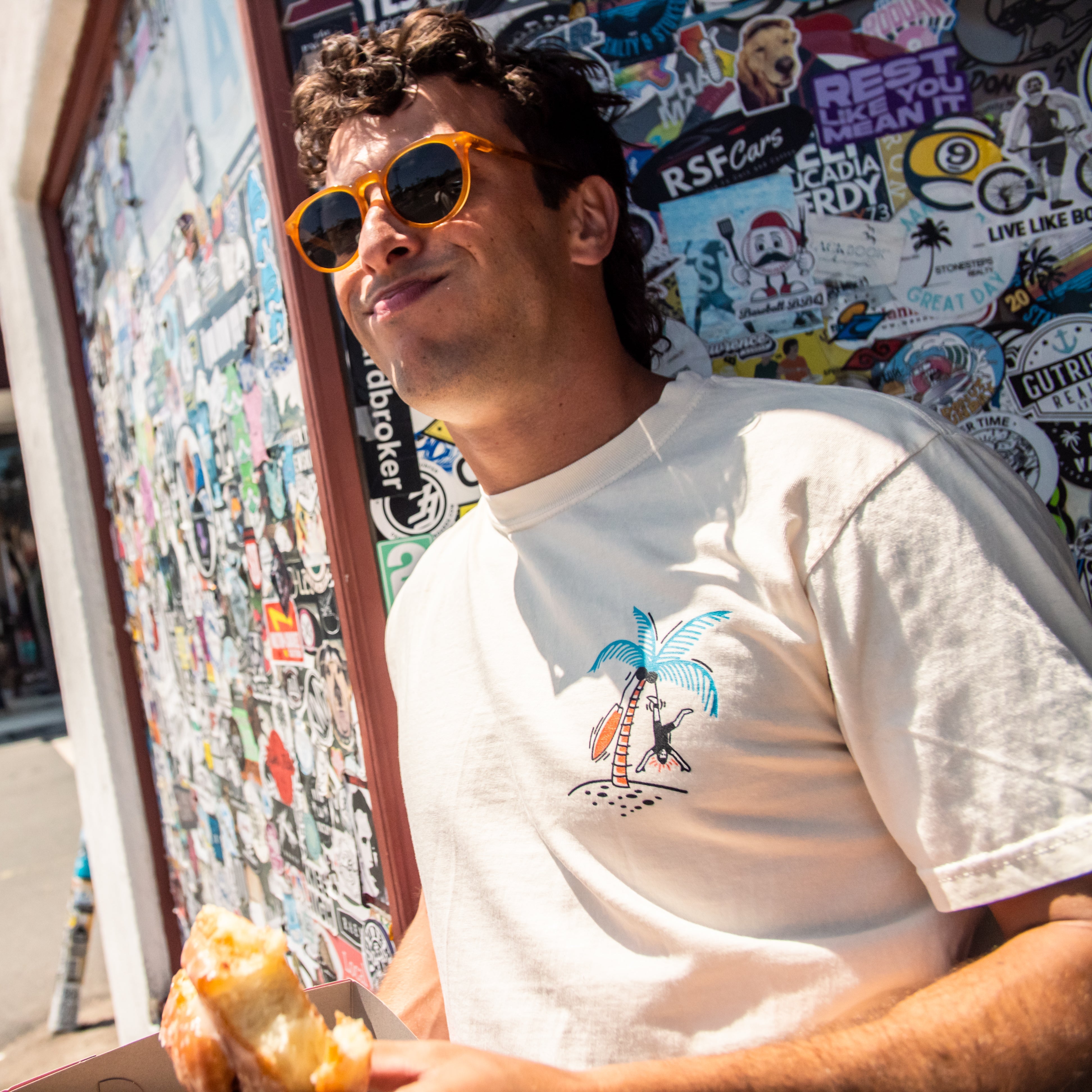 Man wearing sunglasses and a white t-shirt with a graphic design, standing in front of a wall covered in stickers.